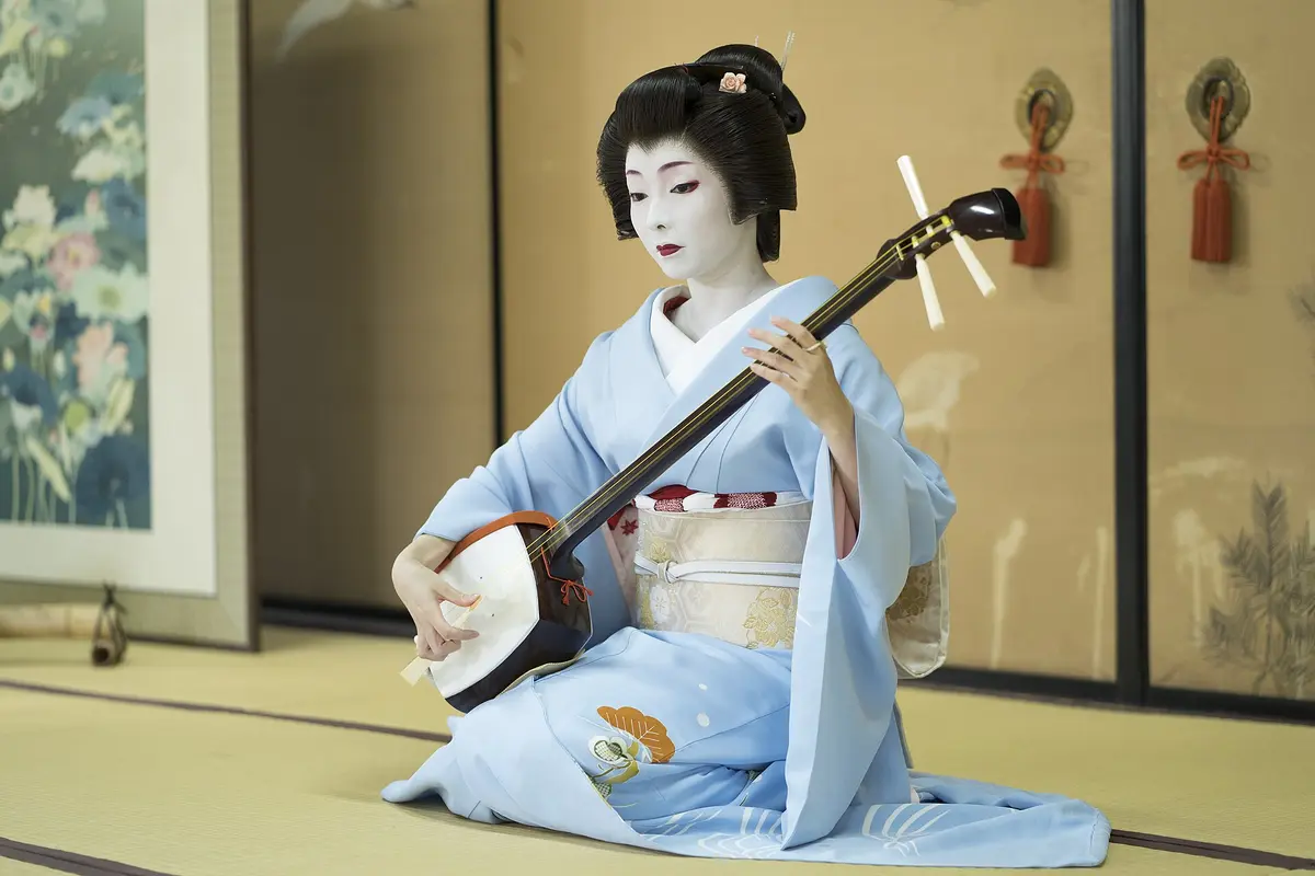 A Kyoto geiko in a pale blue short-sleeved kimono and a wig, kneeling on tatami and playing a shamisen in a temple room