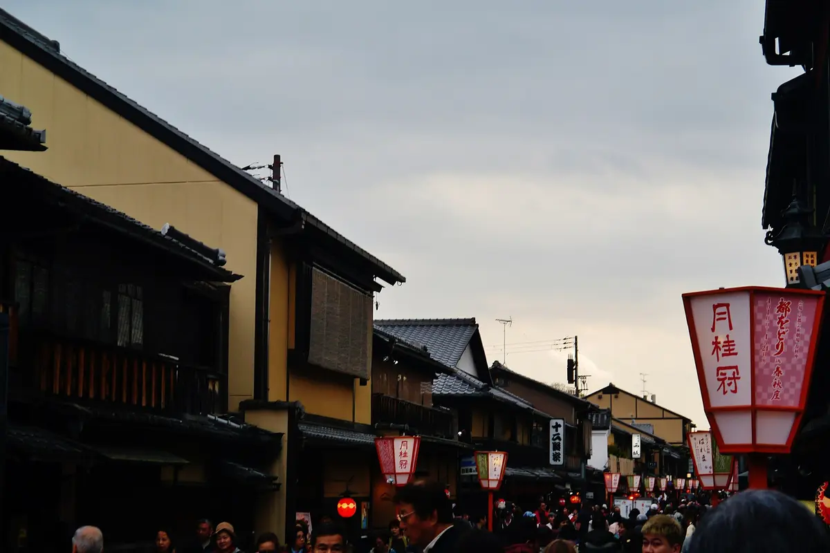 A crowded public street in Gion lined with wooden two-storey townhouses and red and white Miyako Odori lanterns at dusk