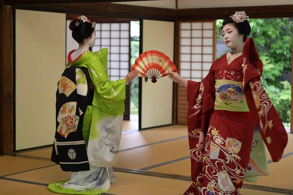 Two maiko dancing with a red and gold fan in a tatami room, one in green with a long black darari obi hanging down her back, one in red